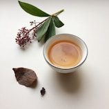 A white teacup filled with light brown tea is placed on a white surface. Accompanying the teacup are a sprig with green leaves and small red berries, a dried brown leaf, and a small piece of dark material, possibly a tea leaf or spice.