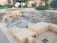A detailed miniature model representing a historical or archaeological site with various structures, including buildings with archways and ruins distributed across a terrain resembling hills. The scene is surrounded by a garden area with trees and shrubs, indicating a carefully designed display.