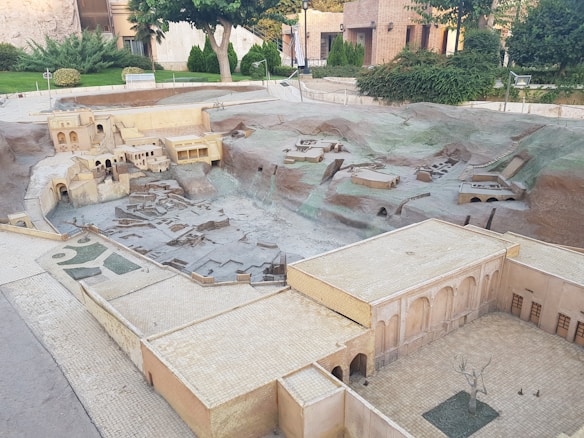 A detailed miniature model representing a historical or archaeological site with various structures, including buildings with archways and ruins distributed across a terrain resembling hills. The scene is surrounded by a garden area with trees and shrubs, indicating a carefully designed display.