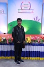 A man stands in front of a colorful display with a banner reading 'Annual Conference 2021' from 'bestelectronics'. The backdrop features a green field design with abstract figures. The area is decorated with a vibrant array of flowers and a draped tablecloth.