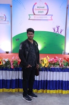 A man stands in front of a colorful display with a banner reading 'Annual Conference 2021' from 'bestelectronics'. The backdrop features a green field design with abstract figures. The area is decorated with a vibrant array of flowers and a draped tablecloth.