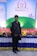 A man stands in front of a colorful display with a banner reading 'Annual Conference 2021' from 'bestelectronics'. The backdrop features a green field design with abstract figures. The area is decorated with a vibrant array of flowers and a draped tablecloth.