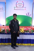 A man stands in front of a colorful display with a banner reading 'Annual Conference 2021' from 'bestelectronics'. The backdrop features a green field design with abstract figures. The area is decorated with a vibrant array of flowers and a draped tablecloth.