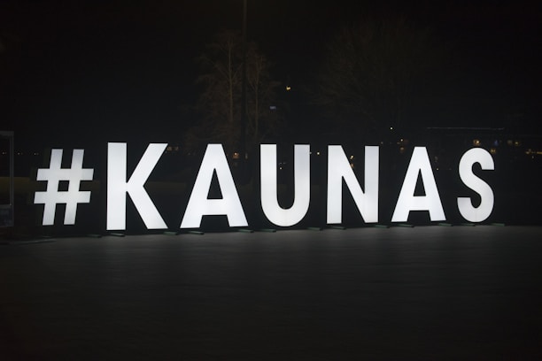 Large illuminated letters spell out #KAUNAS against a dark background, standing on an outdoor pavement area. The letters are evenly spaced and brightly lit, creating a stark contrast with the night around them.