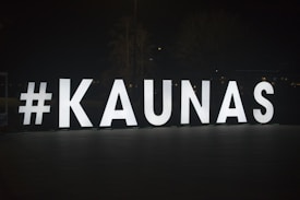 Large illuminated letters spell out #KAUNAS against a dark background, standing on an outdoor pavement area. The letters are evenly spaced and brightly lit, creating a stark contrast with the night around them.