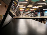 A spacious interior with modern design features an open area with tables and chairs. Colorful geometric ceiling panels and exposed lighting accentuate the industrial look. A few people are visible in the distance, and a tilted screen or partition is in the foreground.