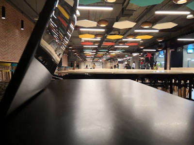 A spacious interior with modern design features an open area with tables and chairs. Colorful geometric ceiling panels and exposed lighting accentuate the industrial look. A few people are visible in the distance, and a tilted screen or partition is in the foreground.