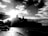 A black and white image depicting a cityscape with a prominent building silhouette against the bright sun. A classic car is parked near a structure labeled 'Self Service', and a sign with 'Cuba' is visible. The sky is filled with scattered clouds, giving a dramatic effect.