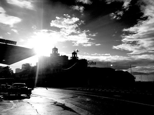 A black and white image depicting a cityscape with a prominent building silhouette against the bright sun. A classic car is parked near a structure labeled 'Self Service', and a sign with 'Cuba' is visible. The sky is filled with scattered clouds, giving a dramatic effect.