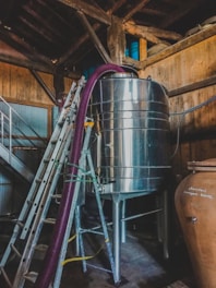 A metallic tank is situated in a wooden structure, with a ladder leaning against it. A purple hose is connected to the tank, and the setting appears industrial or related to beverage production. Wooden beams and barrels are visible in the background.