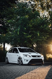 A white, modern hatchback car parked on a textured stone path. It is set against a backdrop of lush green trees, with sunlight filtering through the branches. The car has a sporty design and appears well-maintained, highlighting its sleek curves and shiny metallic rims.