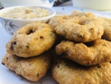 A plate filled with crispy, golden brown fried snacks shaped like donuts is accompanied by a small bowl of creamy beige dip or sauce. The snacks appear to have small green and dark specks, suggesting they contain herbs or spices.