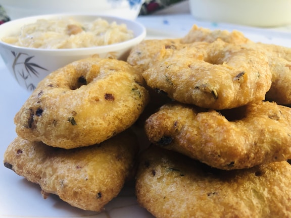 A plate filled with crispy, golden brown fried snacks shaped like donuts is accompanied by a small bowl of creamy beige dip or sauce. The snacks appear to have small green and dark specks, suggesting they contain herbs or spices.