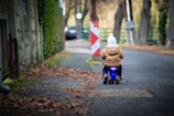 A young child is riding a small blue toy motorcycle down a quiet, tree-lined street. The child is wearing a brown jacket and a white knit hat. The surrounding area has fallen leaves scattered on the ground, indicating it might be autumn. A traffic barrier and a parked car are visible in the background.