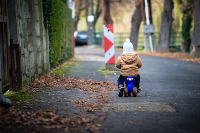 A young child is riding a small blue toy motorcycle down a quiet, tree-lined street. The child is wearing a brown jacket and a white knit hat. The surrounding area has fallen leaves scattered on the ground, indicating it might be autumn. A traffic barrier and a parked car are visible in the background.