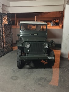A vintage military-style jeep is parked indoors under a structure with concrete floors and white walls. The vehicle has a dark green color and displays a classic, rugged design. Iron grills are visible to the left, with some utilities or equipment seen in the background.