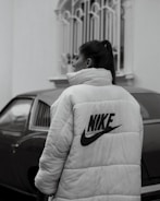 A person stands next to a car, wearing a large puffy jacket with the Nike logo on the back. The scene appears to be outdoors, with a building in the background featuring a decorative window grill.