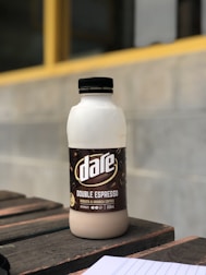 A bottle of 'Dare Double Espresso' is placed on a wooden surface. The label on the bottle displays coffee beans and text indicating it is a robusta and arabica coffee blend. In the background, there is a blurred outdoor wall with a yellow horizontal line.