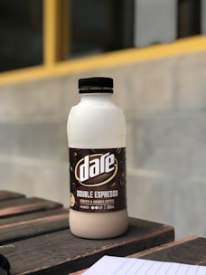 A bottle of 'Dare Double Espresso' is placed on a wooden surface. The label on the bottle displays coffee beans and text indicating it is a robusta and arabica coffee blend. In the background, there is a blurred outdoor wall with a yellow horizontal line.