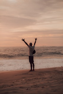 man in white shirt and black shorts standing on seashore during sunset