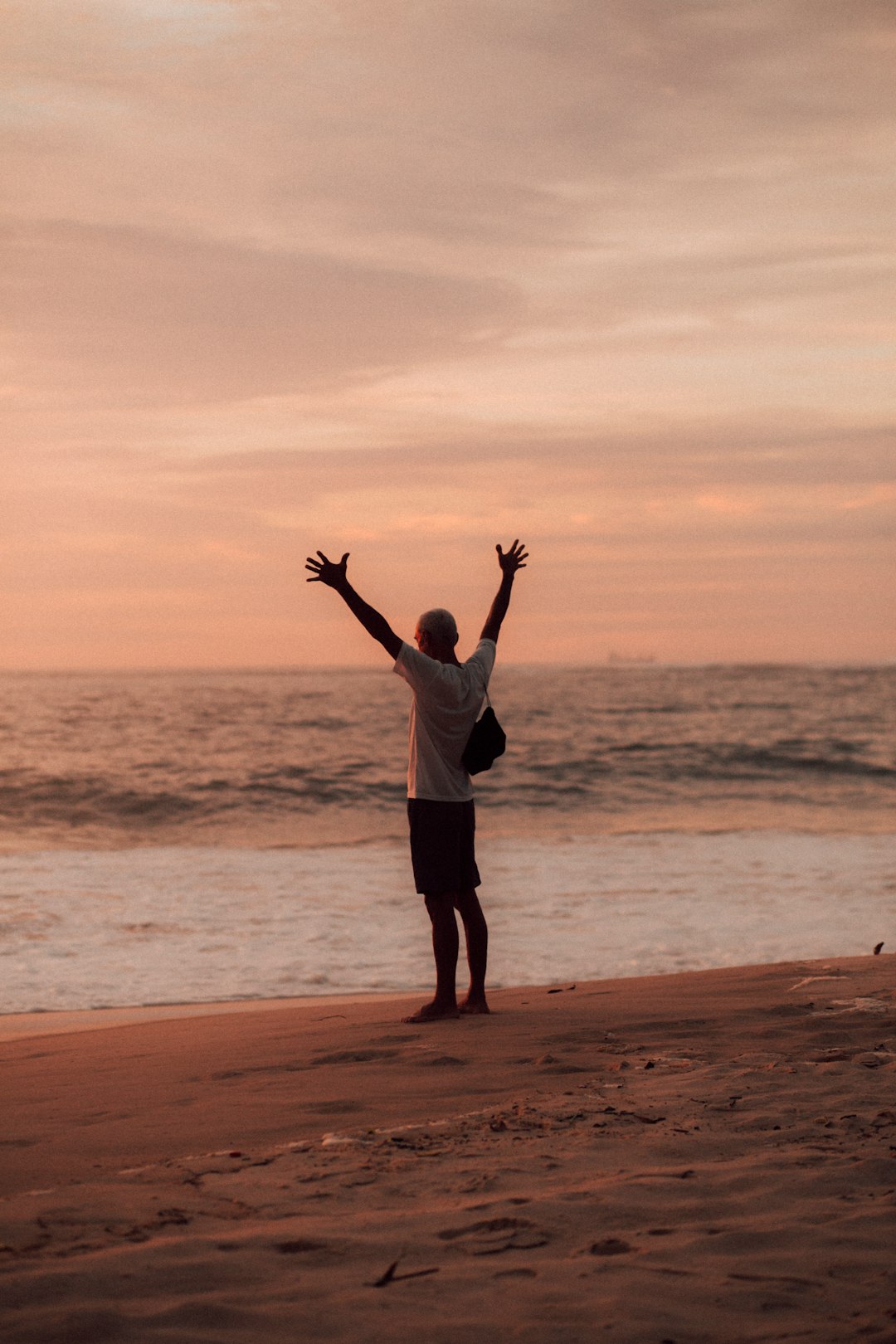 man in white shirt and black shorts standing on seashore during sunset