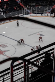 An ice hockey game is in progress inside an indoor arena. The rink has players from two teams wearing contrasting uniforms and referees monitoring the play. Spectators are visible in the stands, and advertisements are displayed along the boards.