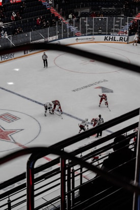 An ice hockey game is in progress inside an indoor arena. The rink has players from two teams wearing contrasting uniforms and referees monitoring the play. Spectators are visible in the stands, and advertisements are displayed along the boards.