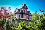 A large, Victorian-style mansion with intricate architectural details stands amidst lush, green foliage. The house features steep, ornate roofs and multiple windows. Crimson and green trees surround the structure, adding a contrasting splash of color beneath a bright blue sky.