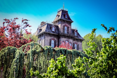 A large, Victorian-style mansion with intricate architectural details stands amidst lush, green foliage. The house features steep, ornate roofs and multiple windows. Crimson and green trees surround the structure, adding a contrasting splash of color beneath a bright blue sky.