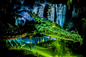 A large, green dragon with textured scales and bright, yellow eyes is situated in a dimly lit cave environment. Its elongated snout and sharp teeth are prominently displayed as it emerges from a pool of water. Chains are visible around the dragon, suggesting captivity. Stalactites hang from the ceiling of the cave, adding to the mysterious atmosphere.