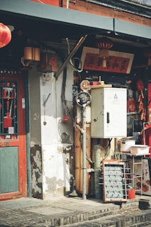 An old storefront with traditional Chinese decorations, including red lanterns and banners with Chinese characters. The building has weathered walls and a combination of modern and vintage elements. There is a foosball table and various utility equipment visible.