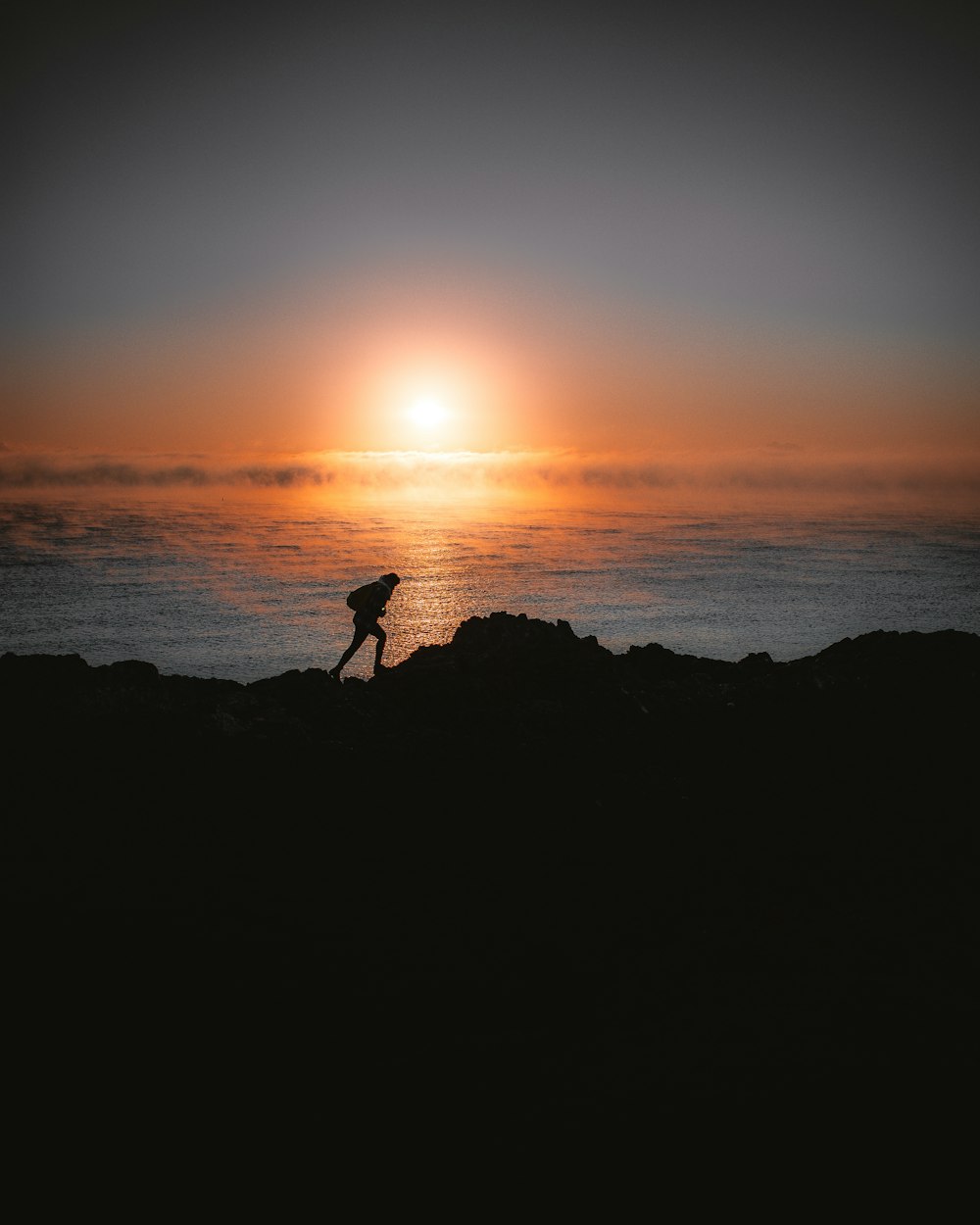 silhouette of person standing on rock near body of water during sunset