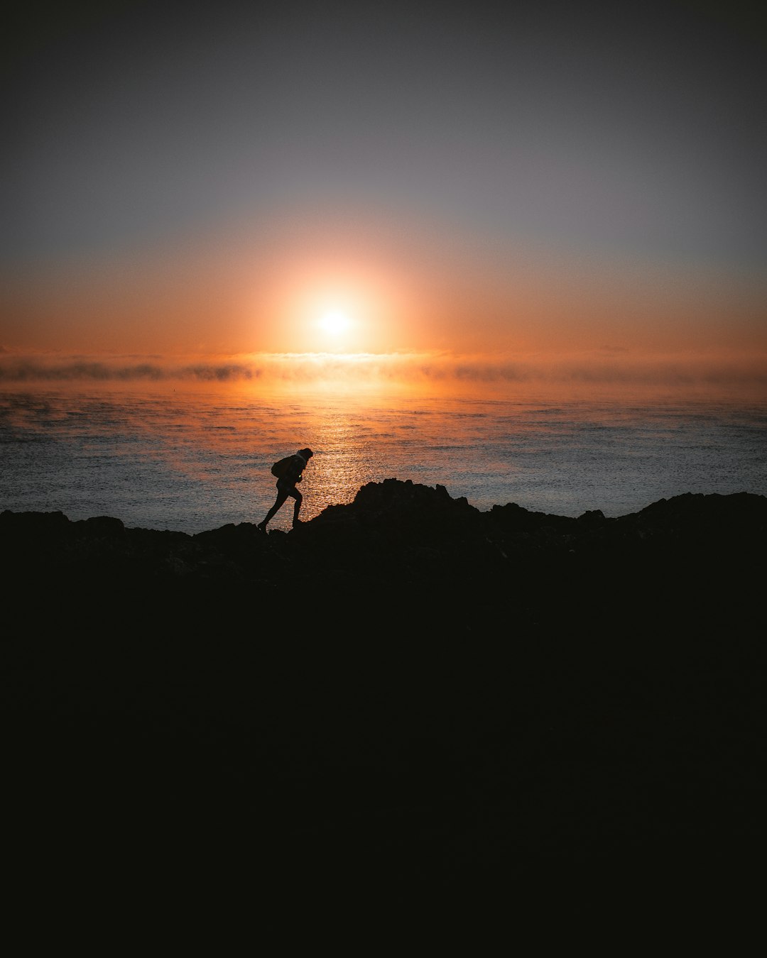 silhouette of person standing on rock near body of water during sunset