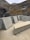 A modern, minimalist concrete structure featuring geometric shapes and clean lines is set against a backdrop of rugged mountain terrain. The landscape includes rocky slopes and a small waterfall meandering through the cliffs.