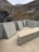 A modern, minimalist concrete structure featuring geometric shapes and clean lines is set against a backdrop of rugged mountain terrain. The landscape includes rocky slopes and a small waterfall meandering through the cliffs.