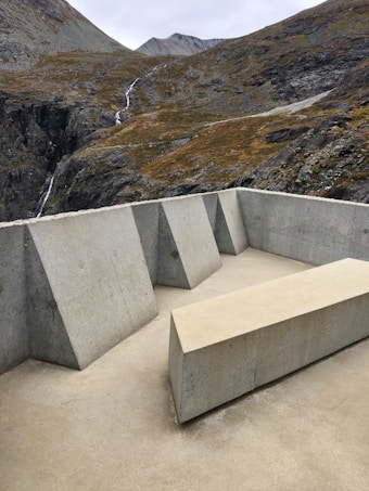 A modern, minimalist concrete structure featuring geometric shapes and clean lines is set against a backdrop of rugged mountain terrain. The landscape includes rocky slopes and a small waterfall meandering through the cliffs.