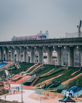 A colorful playground occupies the foreground with vibrant slides and play structures on a sloping green landscape. Overhead, a train is passing on an elevated track with large supporting pillars. The sky is overcast, giving a muted atmosphere.