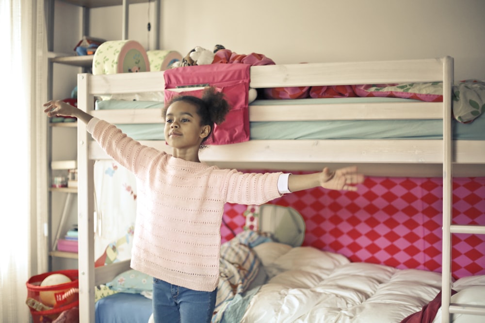 흰색과 빨간색 긴팔 셔츠와 파란색 데님 청바지를 입은 소녀가 침대 근처에 서 있다