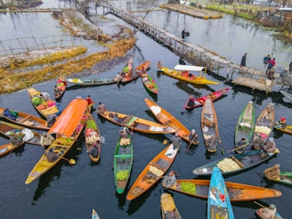 Aerial view of a vibrant floating market featuring multiple wooden boats filled with a variety of goods, including fresh produce. The boats are colorful, and people are engaged in trading activities. A narrow wooden bridge connects small patches of land over the water, with a few people walking on it.