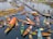 Aerial view of a vibrant floating market featuring multiple wooden boats filled with a variety of goods, including fresh produce. The boats are colorful, and people are engaged in trading activities. A narrow wooden bridge connects small patches of land over the water, with a few people walking on it.