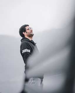 A person with curly hair and glasses is wearing a dark hoodie with the word 'Jordan' on the sleeve. The individual appears to be looking upward and smiling. The background is a gradient of blurred gray and white tones, creating a serene and calm atmosphere.