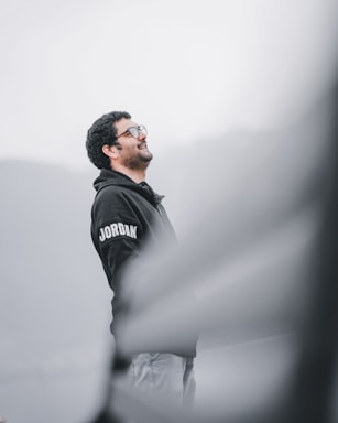A person with curly hair and glasses is wearing a dark hoodie with the word 'Jordan' on the sleeve. The individual appears to be looking upward and smiling. The background is a gradient of blurred gray and white tones, creating a serene and calm atmosphere.