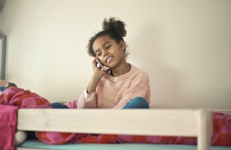 A young girl wearing a pink sweater is sitting on a bed, talking on a mobile phone. The room appears cozy with a colorful blanket draped over the bed.