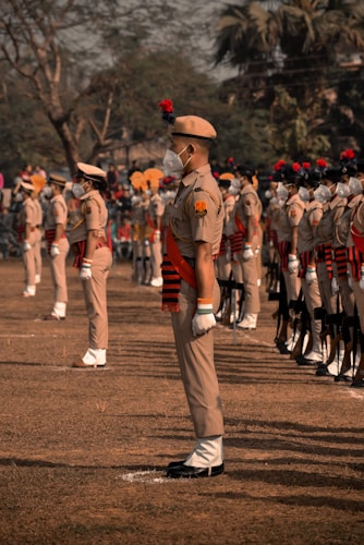 A group of uniformed individuals standing in formation on a grassy field. They are wearing beige uniforms with red and black accents, and some are adorned with red plumes on their hats. All individuals are wearing white masks, and there is a sense of order and discipline in their stance. In the background, there are trees and an audience observing the event.