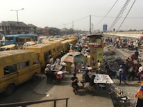 Busy urban street scene with numerous yellow buses lining both sides of the road. A bustling marketplace environment is visible with vendors selling goods under makeshift stalls and umbrellas. People are walking around, engaging with vendors or moving through the area. In the background, there's a view of city buildings and infrastructure like power lines and billboards.