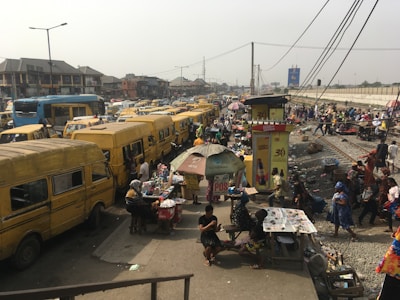 Busy urban street scene with numerous yellow buses lining both sides of the road. A bustling marketplace environment is visible with vendors selling goods under makeshift stalls and umbrellas. People are walking around, engaging with vendors or moving through the area. In the background, there's a view of city buildings and infrastructure like power lines and billboards.