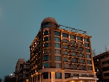 A tall, well-lit hotel building with a modern design stands against a dusky sky. The structure has multiple windows and a prominent signage, suggesting it's a notable establishment. The lighting enhances the warm tones of the building.