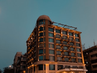 A tall, well-lit hotel building with a modern design stands against a dusky sky. The structure has multiple windows and a prominent signage, suggesting it's a notable establishment. The lighting enhances the warm tones of the building.
