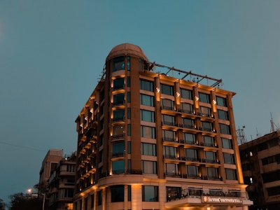 A tall, well-lit hotel building with a modern design stands against a dusky sky. The structure has multiple windows and a prominent signage, suggesting it's a notable establishment. The lighting enhances the warm tones of the building.