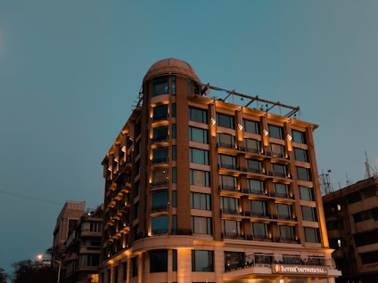 A tall, well-lit hotel building with a modern design stands against a dusky sky. The structure has multiple windows and a prominent signage, suggesting it's a notable establishment. The lighting enhances the warm tones of the building.